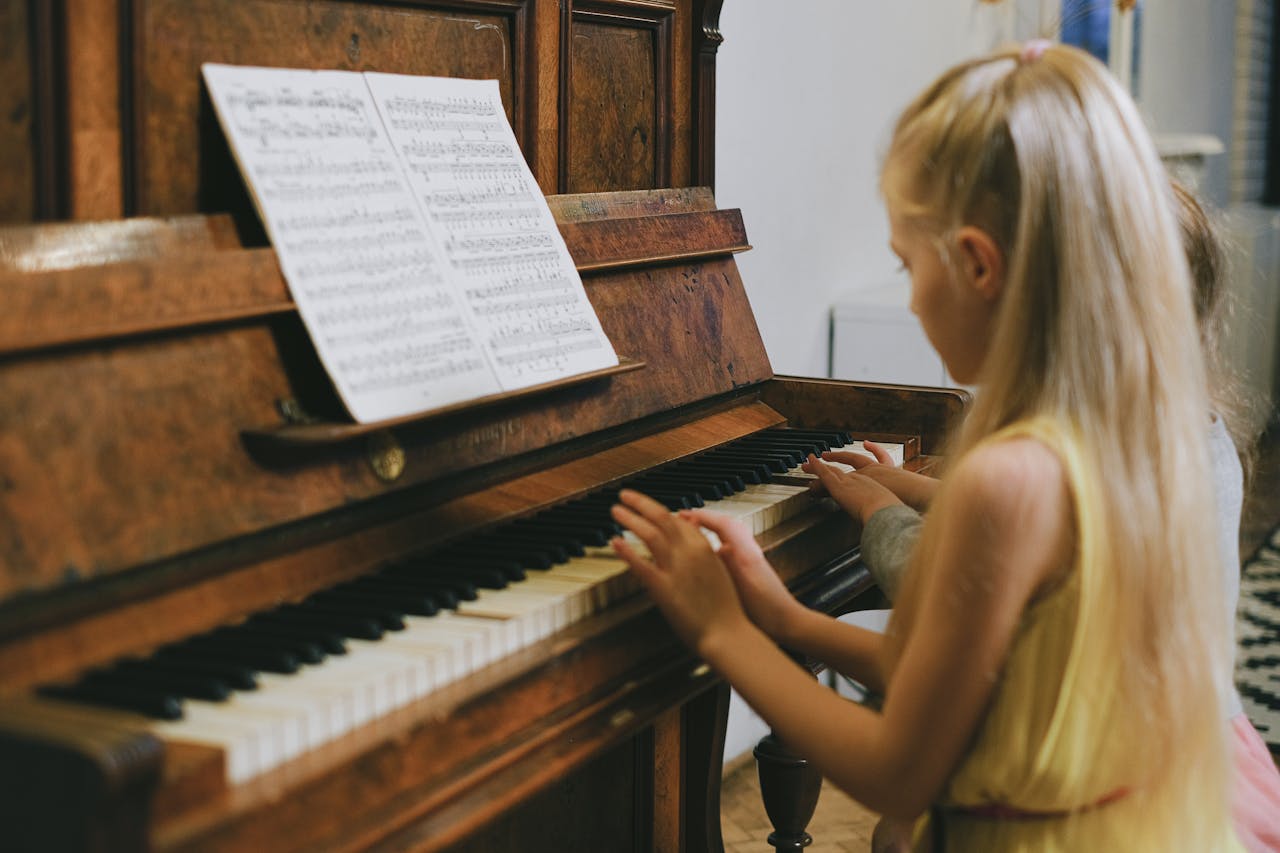 hero-img-02 Two young girls playing piano together indoors, focused on a music sheet.
